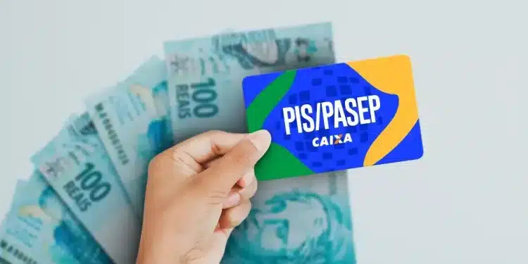 Mão segurando cartão do PIS Pasep da Caixa em frente a cédulas de R$ 100, simbolizando o pagamento do abono salarial em 2026