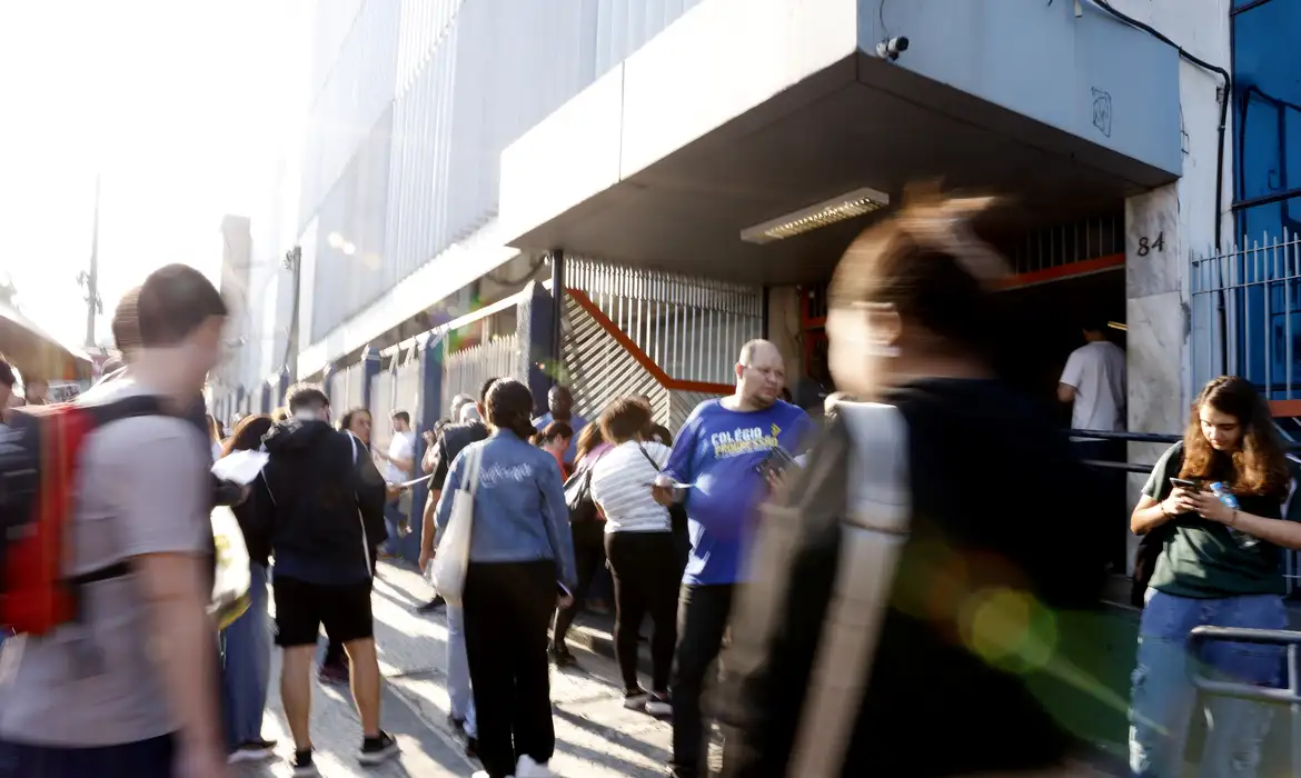 Jovens e adultos aguardam abertura de portão de escola, homem de camisa azul com texto "Colégio" em destaque