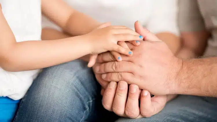 Mãos de criança com esmalte azul entrelaçadas com mãos de adulto em close-up sobre calça jeans.