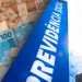 Composição com notas de R$ 50 e R$ 100 espalhadas ao lado da placa azul da Previdência Social fotografada em ângulo inclinado com luz solar ao fundo, ilustrando a antecipação do 13º salário do INSS de 2026, que representa uma injeção de R$ 78,2 bilhões na economia brasileira