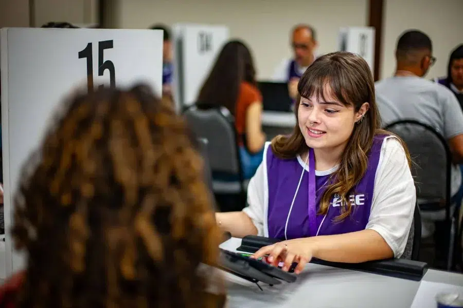 Jovem colaboradora do CIEE com colete roxo atende candidata no guichê 15 durante mutirão de vagas de estágio.