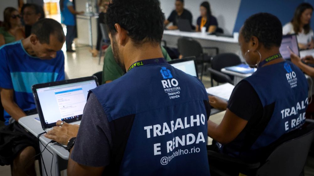 Vista das costas de dois funcionários da Prefeitura do Rio, vestindo coletes azuis com a inscrição "Trabalho e Renda" e o @trabalho.rio. Eles estão sentados à mesa operando notebooks para cadastrar cidadãos que aparecem sentados à frente deles em uma sala de atendimento.