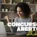 Mulher negra de cabelos cacheados sorri e comemora com os braços levantados em frente a um notebook aberto, com os textos "Concursos Abertos 2026" sobrepostos em branco e amarelo, representando candidatos que podem se inscrever no concurso público da Prefeitura Municipal de Guzolândia — SP, com vagas para Escriturário, Nutricionista Clínico, Médico Pediatra e outros cargos em 2026