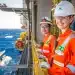 Funcionárias da Petrobras sorrindo em plataforma offshore, com uniforme de segurança laranja, em ambiente marítimo.