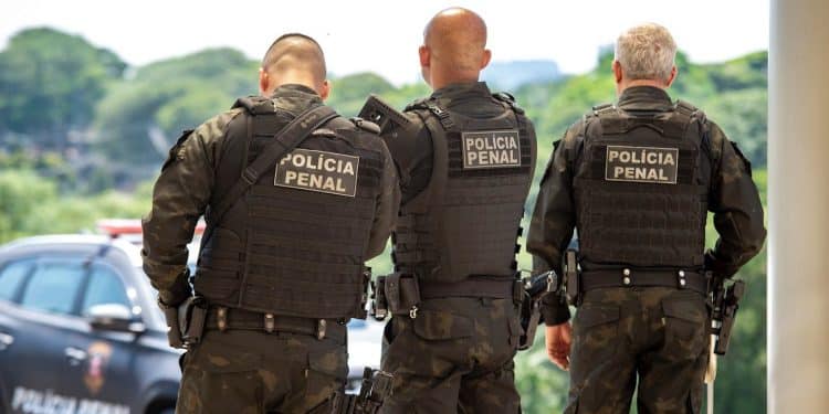 Três policiais penais de costas usando coletes pretos com inscrição "Polícia Penal" e viatura estacionada