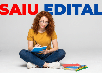 Jovem ruiva de óculos sentada no chão, escrevendo em caderno azul, com pilha de cadernos coloridos ao lado e texto "Saiu Edital"