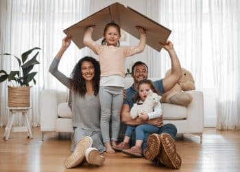 Pais e filhos brincando com caixa de papelão simulando casa em sala de estar.