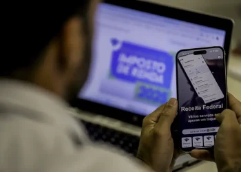 Homem acessa serviços da Receita Federal pelo celular enquanto visualiza portal do Imposto de Renda no notebook