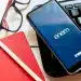 Celular com o aplicativo do Enem sobre um caderno vermelho, lápis, óculos e teclado representando a preparação para o Enem 2026.