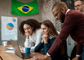 Equipe diversificada de jovens profissionais reunida em torno de um notebook em ambiente corporativo com bandeira do Brasil ao fundo