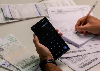 Consumidor fazendo contas em calculadora do celular cercado de faturas de energia elétrica com suspeita de empréstimo indevido
