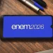 Tela de smartphone com o logo do Enem 2026 em fundo azul, com canetas e cadernos ao redor.
