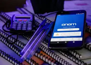 Celular com página do Enem aberta sobre cadernos, canetas e calculadora, representando o processo de solicitação de isenção da taxa do Enem 2026 e a justificativa de ausência