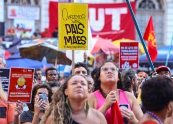 Multidão em ato sindical com cartaz amarelo "Queremos ser pais e mães presentes" e bandeiras da CUT