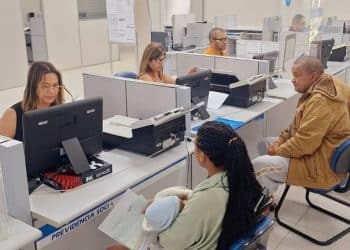 Servidores do INSS atendendo segurados em balcões com computadores em agência da Previdência Social, com placas indicativas e cadeiras para o público