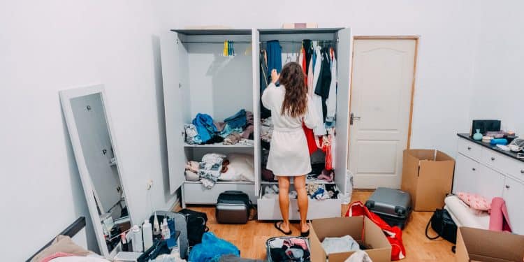 Acumulação de coisas psicologia - mulher de costas diante de guarda-roupa lotado com roupas e objetos espalhados pelo quarto