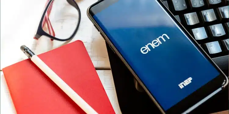 Imagem mostrando o celular com o logo do Enem 2026 na tela, com caderno, caneta e óculos sobre a mesa. A tela do celular exibe o aplicativo do Enem.