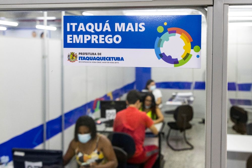 Vista através de um vidro de uma unidade de atendimento do Itaquá Mais Emprego. No vidro, há um adesivo azul e branco com o logotipo do programa e a inscrição "Prefeitura de Itaquaquecetuba". Ao fundo, de forma levemente desfocada, veem-se pessoas sentadas em mesas de atendimento em um escritório organizado.