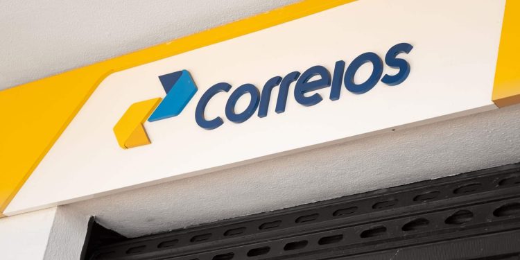 Placa branca com logo dos Correios em azul e amarelo na fachada da agência, com grade de ferro.