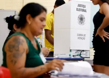 Mesária e eleitores em cabine de votação durante as eleições, destacando os mesários no processo eleitoral.