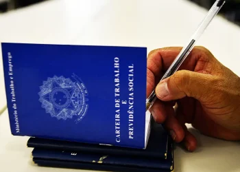 Mão segurando caneta sobre carteiras de trabalho azuis em ilustração sobre a aprovação do piso salarial nacional dos assistentes sociais no Brasil