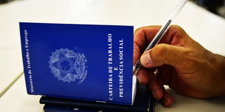 Mão segurando caneta sobre carteiras de trabalho azuis em ilustração sobre a aprovação do piso salarial nacional dos assistentes sociais no Brasil