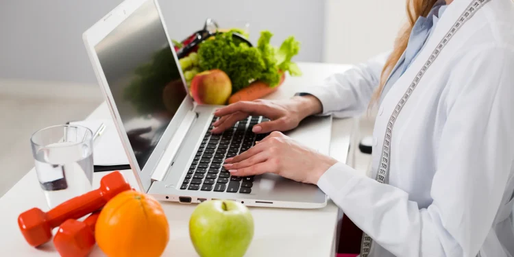 Estudante de Nutrição analisando alimentos saudáveis enquanto utiliza notebook em ambiente de estudo.