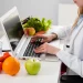 Estudante de Nutrição analisando alimentos saudáveis enquanto utiliza notebook em ambiente de estudo.