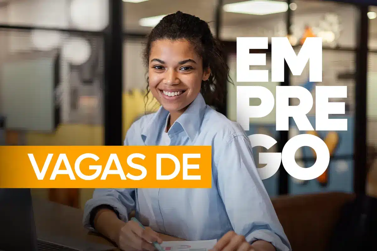 Mulher com cabelos cacheados e óculos sorri enquanto digita em um notebook prata. Ela está em uma sala iluminada com decoração moderna, flores amarelas em um vaso à esquerda e uma caneca verde sobre a mesa branca.