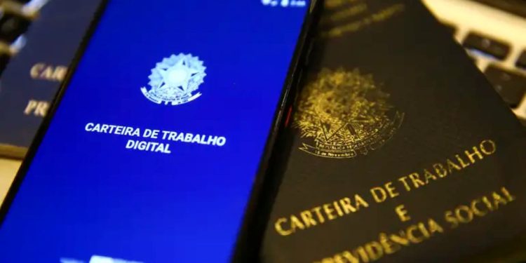 Close-up mostrando um smartphone com a tela azul ligada no aplicativo da Carteira de Trabalho Digital, posicionado ao lado de uma Carteira de Trabalho física (caderneta preta tradicional) com o brasão da República em dourado. O fundo é levemente desfocado, sugerindo um ambiente de escritório ou busca por emprego.