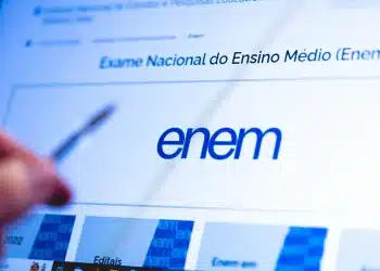 Mão segurando caneta apontada para tela de computador exibindo a página oficial do Inep com o título "Exame Nacional do Ensino Médio (Enem)" e o logo azul do exame em destaque, representando as orientações e critérios de correção da redação do Enem 2026