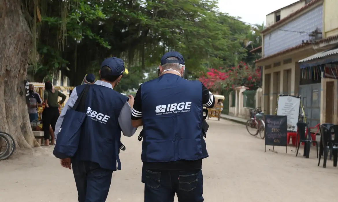 Dois agentes do IBGE de costas, usando coletes com a identificação do instituto, caminhando em área urbana, representando os profissionais que atuam nos cargos de Agente de Pesquisas e Mapeamento e Supervisor de Coleta e Qualidade