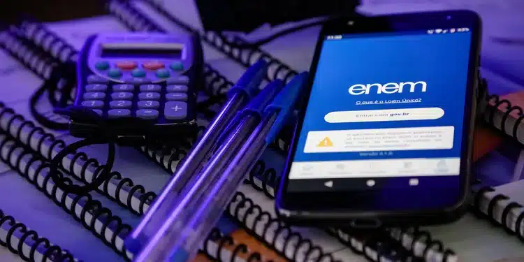 Estudante se preparando para o Enem com celular aberto no aplicativo, cadernos, canetas e calculadora sobre a mesa.