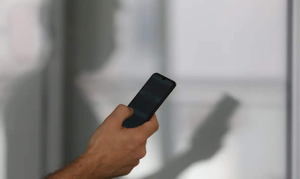 Homem segurando celular escuro ao lado da sombra projetada na parede em ilustração sobre o direito à desconexão do trabalhador fora do expediente