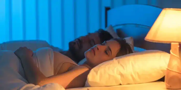 Casal dormindo com a luz acesa no quarto, ambiente com iluminação suave e azulada, refletindo os efeitos do sono com luz.