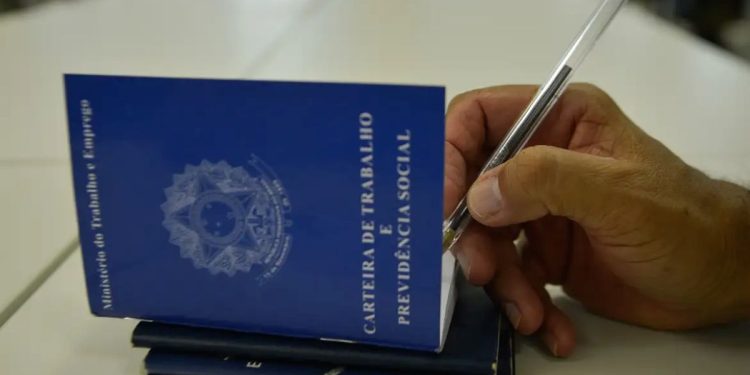 Close de uma mão masculina segurando uma caneta sobre uma Carteira de Trabalho e Previdência Social (CTPS) azul aberta. A mão está prestes a assinar ou preencher o documento sobre uma mesa branca.