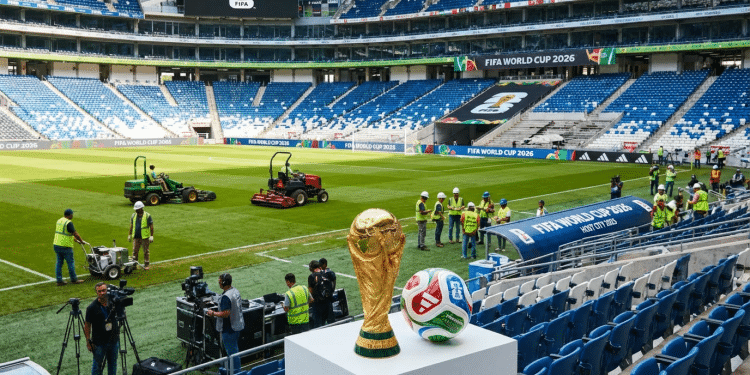 Troféu dourado da Copa do Mundo FIFA 2026 e bola oficial em pedestal branco com trabalhadores no gramado do estádio ao fundo