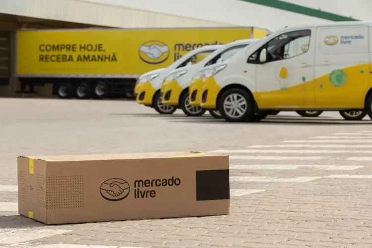 Caixa do Mercado Livre em piso de paralelepípedo, com vans e caminhão amarelo da frota ao fundo no CD.