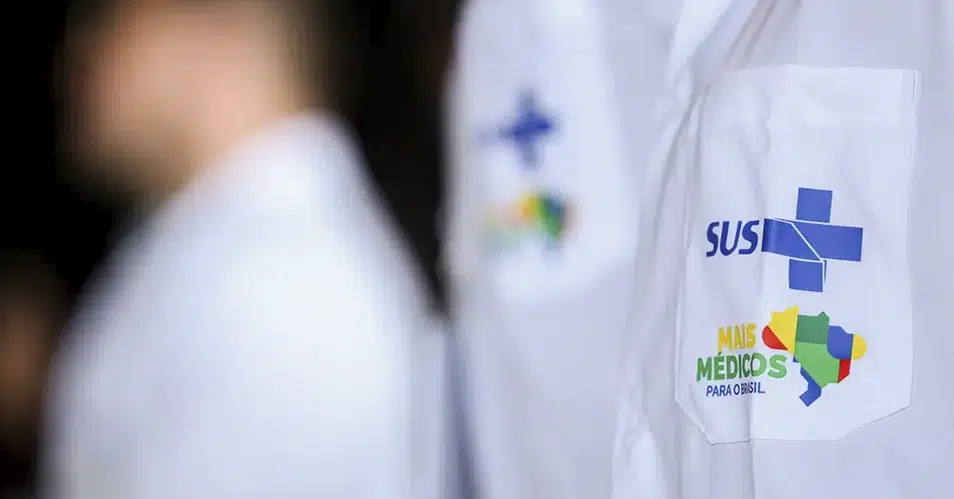 Close lateral de um jaleco branco de profissional de saúde. No bolso frontal, aparecem bordados os logotipos do SUS (Sistema Único de Saúde) em azul e do programa Mais Médicos para o Brasil em verde, laranja e azul, acompanhados de um mapa do Brasil estilizado e colorido. O fundo mostra outros profissionais em desfoque.