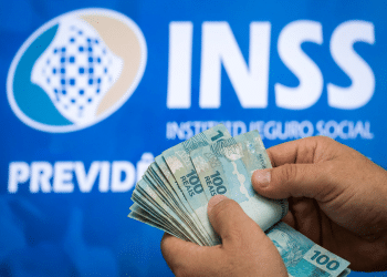 Mãos contando notas de R$ 100 com logo do INSS e o texto Previdência Social ao fundo, em painel azul, simbolizando o pagamento da segunda parcela do 13º salário e os golpes a ela relacionados.