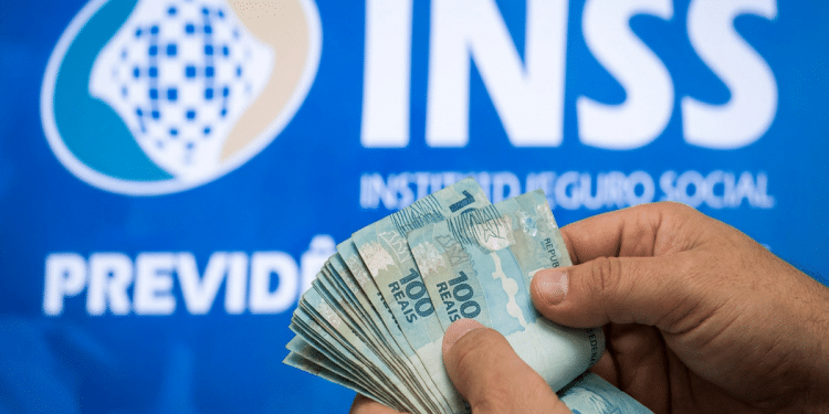 Mãos contando notas de R$ 100 com logo do INSS e o texto Previdência Social ao fundo, em painel azul, simbolizando o pagamento da segunda parcela do 13º salário e os golpes a ela relacionados.