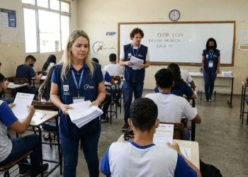 Aplicadores com coletes azuis distribuem provas para estudantes em sala de aula do Enem.