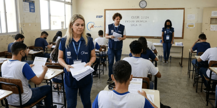 Aplicadores com coletes azuis distribuem provas para estudantes em sala de aula do Enem.