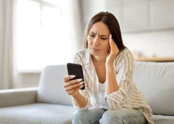 Mulher preocupada olha celular sentada no sofá de casa.