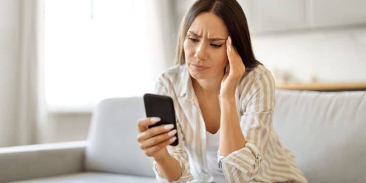 Mulher preocupada olha celular sentada no sofá de casa.