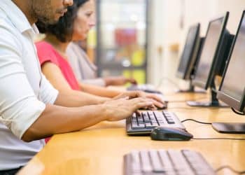 Três pessoas sentadas em fila digitando em teclados pretos diante de monitores em mesa de madeira, simbolizando os cursos livres.