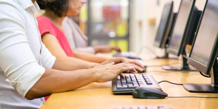 Três pessoas sentadas em fila digitando em teclados pretos diante de monitores em mesa de madeira, simbolizando os cursos livres.