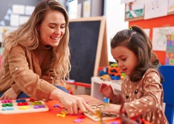 Mulher loira sorridente ensina menina a encaixar letras coloridas em tabuleiro numa sala de aula infantil.