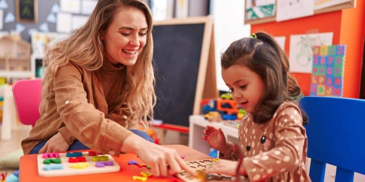 Mulher loira sorridente ensina menina a encaixar letras coloridas em tabuleiro numa sala de aula infantil.