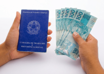 Mãos segurando carteira de trabalho e notas de 100 reais.